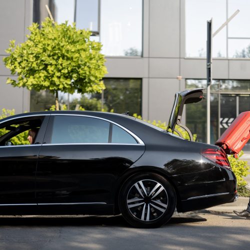 Chauffeur of a business-class car packs red suitcase into a trunk on the street near an office building. Concept of business trips, personal driver or luxury taxi